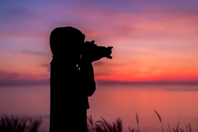 Silhouette woman photographing sea against sky during sunset