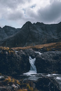 Scenic view of waterfall by mountains against cloudy sky