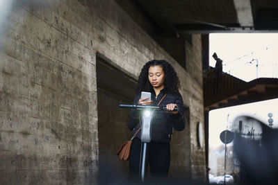 Teenage girl with electric push scooter using smart phone below bridge