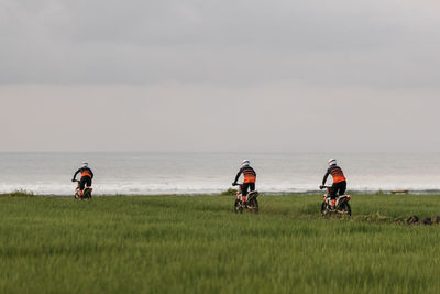 People riding dirt bikes on grass by sea against sky