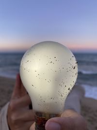 Cropped hand of person holding light bulb at beach against sky