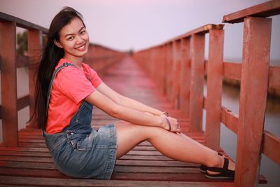 Side view portrait of smiling young woman sitting on pier during sunset