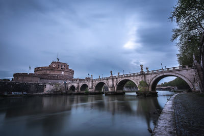 Arch bridge over river by buildings in city during sunset