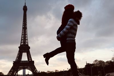 Man lifting woman while embracing against eiffel tower and cloudy sky