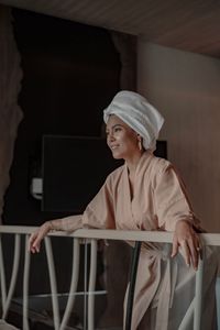 Smiling woman wearing bathrobe and looking away while standing by railing at home