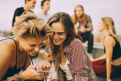 Smiling woman showing mobile phone to friend while people enjoying at building terrace during party