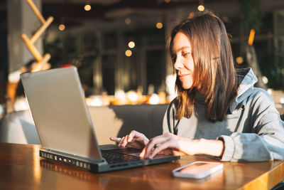 Young brunette woman in casual clothes working with laptop using mobile phone at cafe