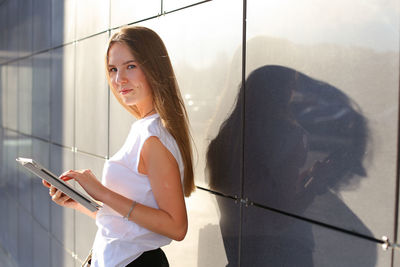 Young woman using digital tablet standing by wall