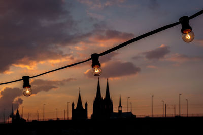 Low angle view of illuminated light bulbs by silhouette church against sky during sunset