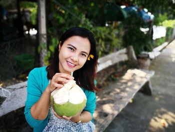 Portrait of smiling woman having coconut water while sitting on bench at footpath