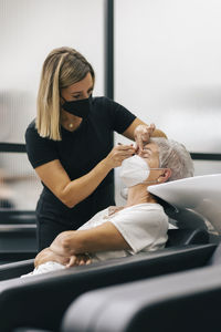 Female beautician with face mask tweezing customer's eyebrows in beauty spa