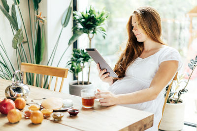A pregnant woman is eating breakfast at the table and looking into a tablet. communication