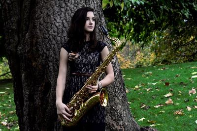 Woman holding saxophone while standing against tree