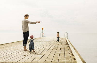 Mother showing something to baby girl while standing on pier at sea against sky