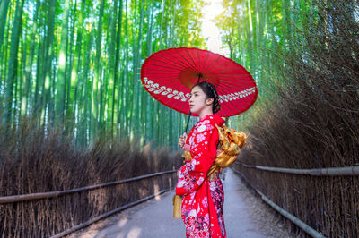 Mid adult woman holding red umbrella on footpath amidst bamboo forest