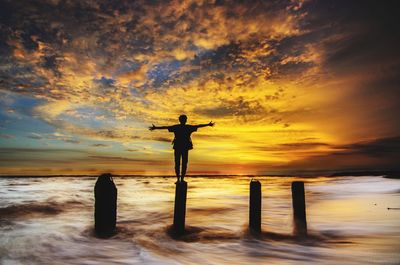 Silhouette man with arms outstretched standing on wooden post in sea against orange sky