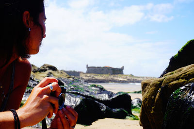 Woman holding camera sitting by rock formation against sky