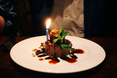 Close-up of illuminated candle on dessert in plate over table with man sitting in background