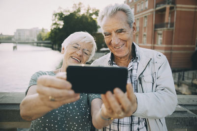 Smiling senior couple taking selfie with mobile phone while standing against railing in city