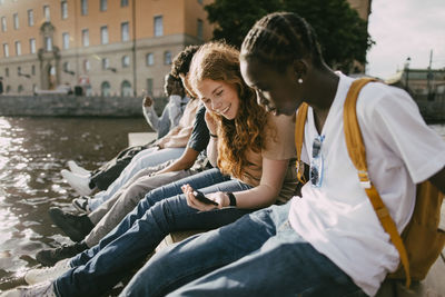 Smiling teenage girl sharing smart phone with male friend while sitting on pier near lake