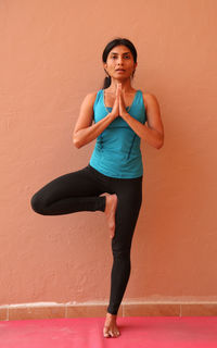 Full length of woman performing yoga while standing against orange wall