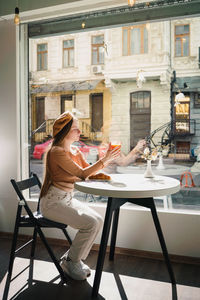 French female in beret sitting at table in cafe with aromatic glass of coffee and freshly baked croissant