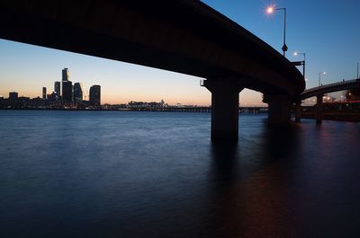 Illuminated bridge over river by buildings against sky during sunset