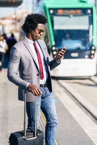 Businessman with luggage using phone while standing in city