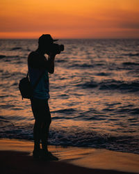 Side view of silhouette man photographing on shore at beach during sunset