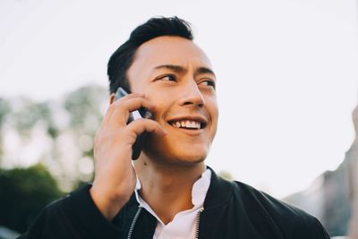 Smiling man talking through smart phone while looking away against sky