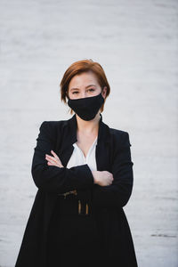 Determined female entrepreneur wearing elegant suit and protective mask standing with crossed arms near building and looking at camera