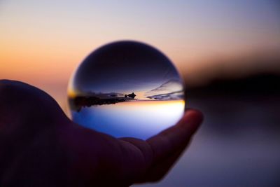 Close-up of hand holding crystal ball against sky during sunset