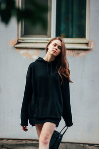 Portrait of young woman standing on footpath against wall