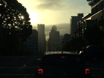 Traffic on road by buildings against sky during sunset