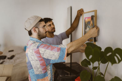 Side view of man hanging frame on wall with boyfriend at home