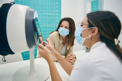 Side view of cosmetologist and customer sitting at table with facial skin diagnostics equipment while looking at tablet and discussing results of analysis in beauty clinic