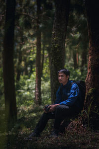 Young man looking away while sitting on tree trunk in forest