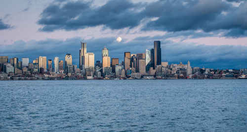 Cityscape by river against cloudy sky at dusk
