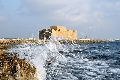 Waves splashing on rocky shore by castle against sky