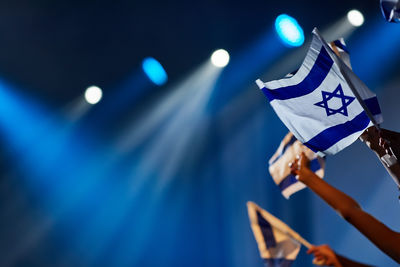 Hands at a concert hold the flag of israel against the background of blue rays of lighting