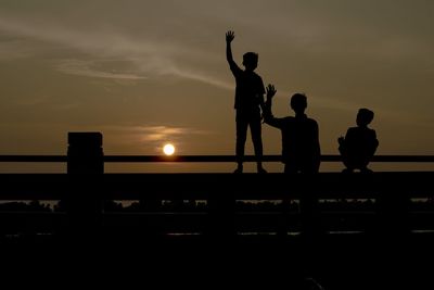 Silhouette male friends on railing against sky during sunset