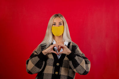 Portrait of woman wearing mask making heart shape against red background