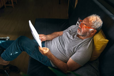 Handsome mature man wearing reading glasses sitting on sofa looks at documents while works at home. 