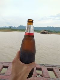 Midsection of person holding beer bottle against sky