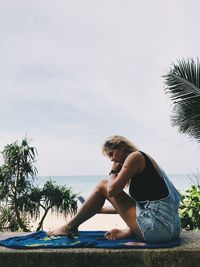 Young woman sitting on palm tree by sea against sky