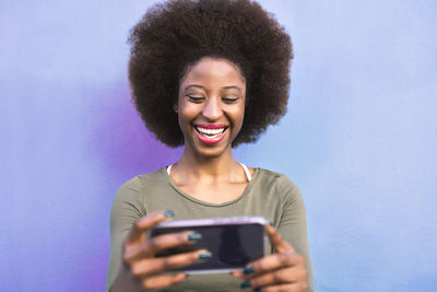 Cheerful young woman using smart phone while standing against wall