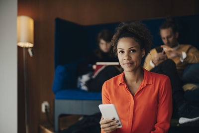 Portrait of confident businesswoman using smart phone while sitting against colleagues at office in cafeteria