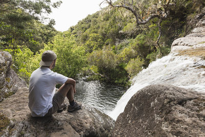 Rear view of man sitting on rock looking at waterfall in madeira island. 