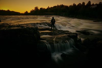 Silhouette man standing by waterfall against sky during sunset