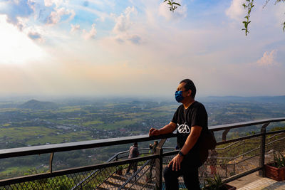 Rear view of man standing by railing against sky during sunset